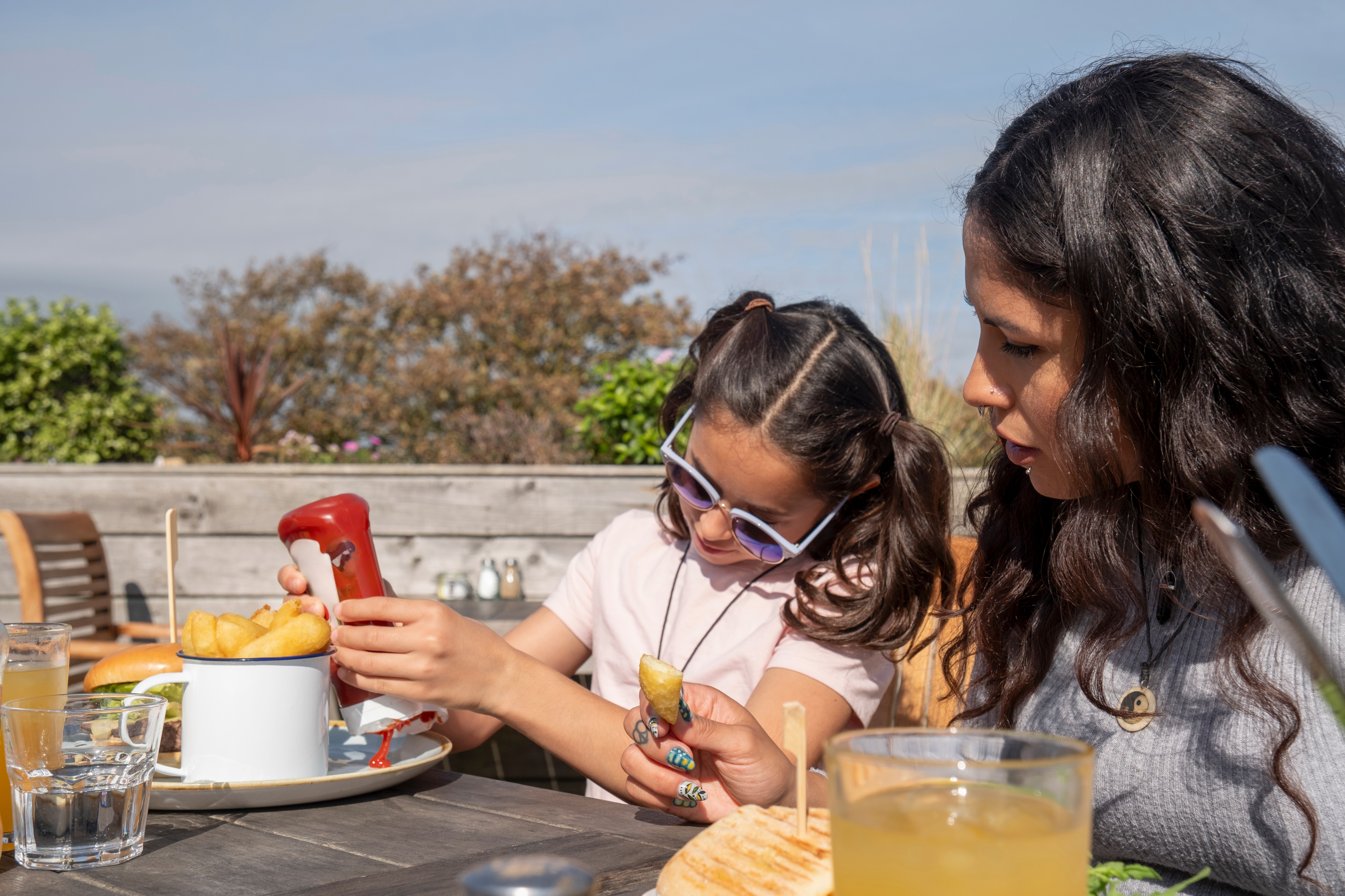 Female with dark hair looks on at younger girl squeezing ketchup onto a plate of chips, outdoors