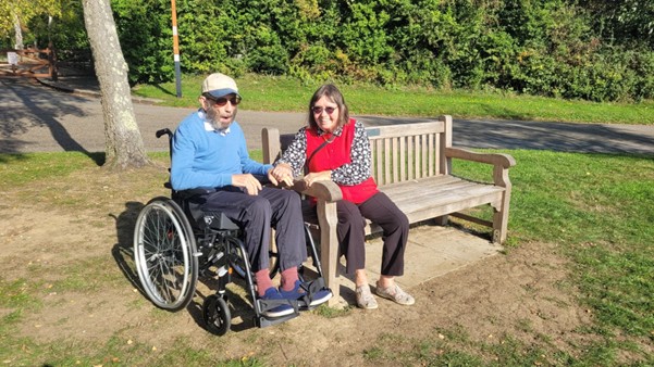Pat sits on a bench in a park, holding the hand of her husband Geoff, who is using a wheelchair